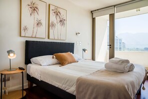 A neatly made bed with white linens and a brown pillow, a bedside lamp, and a window with a view of buildings and mountains.