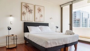 Une chambre moderne avec un lit, deux lampes fixées au mur et une grande fenêtre donnant sur la ville.