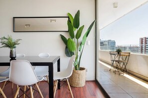 A modern room with a dining table, chairs, a large potted plant, and a view of the city through a large window. - Modern 2-Bedroom, 2-Bath Apartment For 5 Guests (Las Condes)
