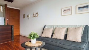 A living room with a dark grey sofa, a small round table with a plant, and a woven placemat. - Cozy 1BR in Prime Location: Amenities & City Views (Las Condes)