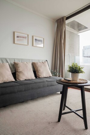Ein modernes Wohnzimmer mit einem grauen Sofa, zwei gerahmten Bildern, einer Zimmerpflanze und Blick durch ein großes Fenster.