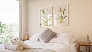 Un lit bien fait, avec une couette blanche, un oreiller gris et une serviette enroulée posée sur le côté. Deux reproductions encadrées de motifs botaniques sont accrochées au mur, au-dessus du lit.