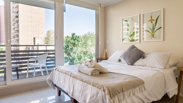 Une chambre à coucher comprenant un lit, une table de chevet, une fenêtre donnant sur un immeuble et des arbres, ainsi que deux reproductions botaniques encadrées accrochées au mur.
