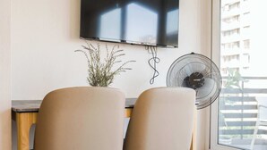 Une pièce équipée d’un téléviseur à écran plat fixé au mur, d’un ventilateur sur pied, de deux chaises et d’une plante en pot.