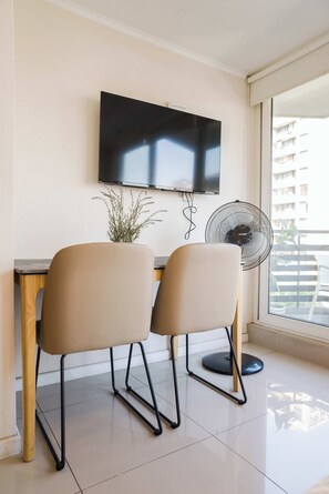 Una habitación con una televisión de pantalla plana montada en la pared, un ventilador de pie, dos sillas y una planta en maceta.