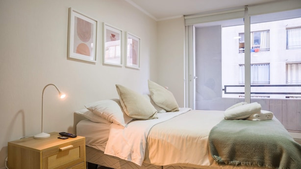 A bedroom with a bed, bedside table, lamp, and artwork on the wall. - Downtown Stgo Studio: Balcony, WiFi & City Views! (Santiago)