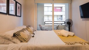 Une chambre d’hôtel équipée d’un lit, d’un ventilateur, d’une télévision et d’un balcon donnant sur des immeubles.