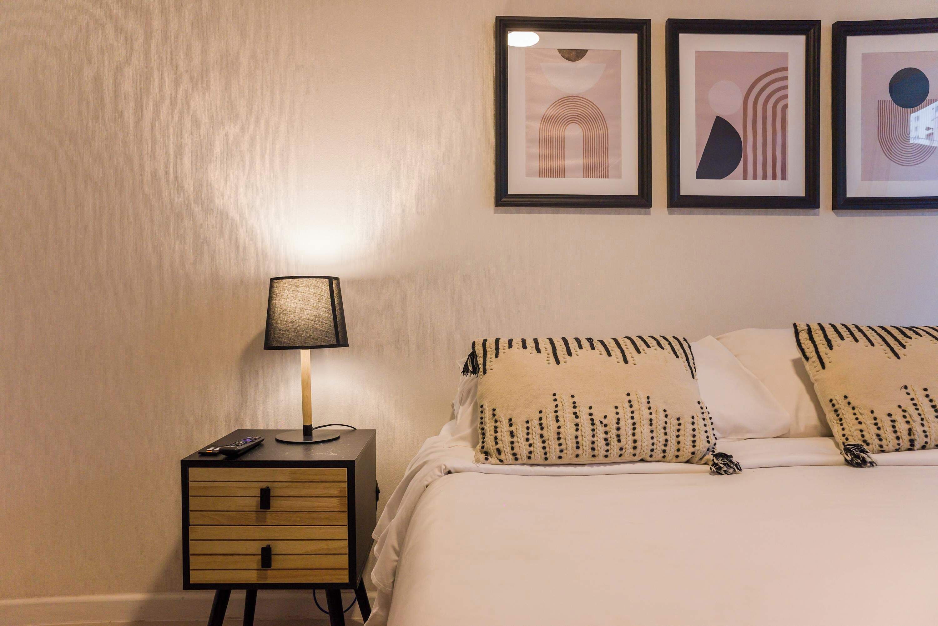 Un lit bien fait, agrémenté d’oreillers à motifs, une lampe de chevet, une petite table de nuit en bois avec une télécommande, et trois œuvres d’art encadrées accrochées au mur.