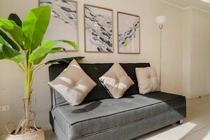 A grey sofa with three beige cushions, a large green plant in a woven basket, and three abstract wall art pieces above.