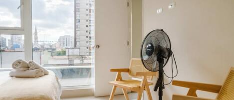 Hotelkamer met een bed, een ventilator, twee houten stoelen en een groot raam met uitzicht op de stad.