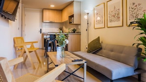 Un salon moderne avec un canapé, une table basse et un téléviseur. On aperçoit à l’arrière-plan un coin cuisine avec des meubles en bois et une table à manger.