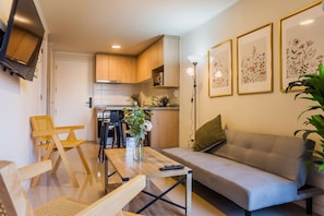 Una sala moderna con un sofá, una mesa de centro y un televisor. Se aprecia una cocina al fondo, con armarios de madera y una mesa de comedor.