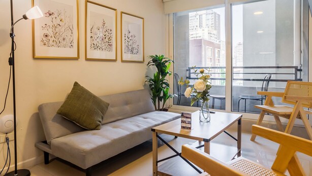 Un salon moderne avec un canapé, une table basse et un coin repas. On peut y admirer des œuvres d'art florales encadrées au mur, une plante en pot et une vue sur la ville à travers de grandes fenêtres.