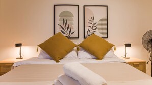 Un lit bien fait, avec des draps blancs, deux coussins de couleur moutarde et une serviette blanche enroulée. Au-dessus du lit, on peut voir deux œuvres d'art abstraites encadrées.