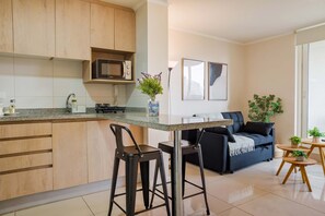 A modern kitchen with wooden cabinets, a microwave, and a bar stool. There is a sofa and a small table with plants in the living area.