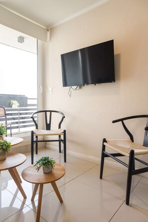 A modern room with a flat-screen TV mounted on the wall, two wooden chairs with woven seats, a small round table, and a potted plant.