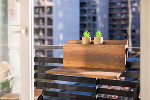 Un balcón con un banco de madera, dos macetas con plantas y vistas a edificios altos.