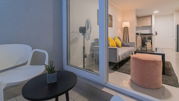 Een moderne woonkamer met een glazen deur die leidt naar een keuken, een eethoek en een klein balkon met een tafel en stoelen.