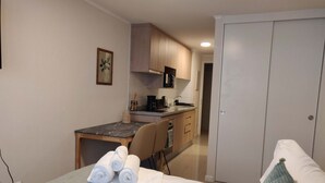 Une chambre avec un lit, une table de chevet avec des serviettes, un petit bureau, une chaise et, au fond, une kitchenette.