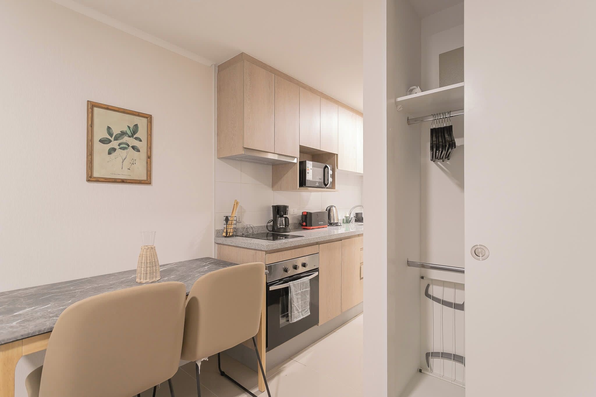 Cocina compacta con microondas, cafetera y horno pequeño. Hay una mesa de comedor con sillas e ilustración botánica enmarcada en la pared.