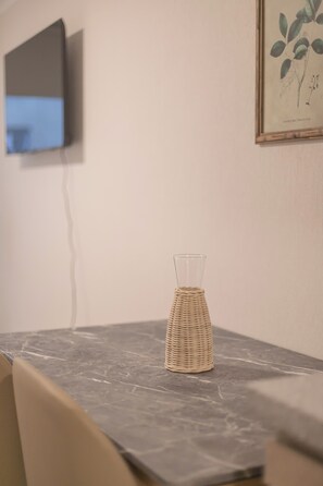 Une pièce comprenant une table en marbre, un vase en osier, une estampe botanique encadrée et un téléviseur fixé au mur.