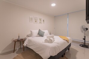 Une chambre à coucher comprenant un lit, une table de chevet, un ventilateur sur pied et des œuvres d’art botanique encadrées accrochées au mur.