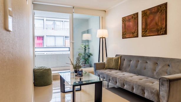 A modern living room with a glass coffee table, a grey sofa, and two wall-mounted lamps. - Santiago: 1BR Apartment with Balcony and Views (Santiago)