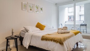 A hotel room with a bed, a small table with a lamp, a balcony with chairs, and framed botanical prints on the wall. - Santiago: 1BR Apartment with Balcony and Views (Santiago)