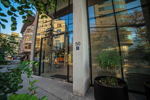 Modern glass-fronted building entrance at Avenida 50, framed by leafy planters and a lively urban streetscape. - Functional and Bright Apartment in Providencia (Santiago)