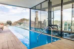 Una piscina en la azotea con paredes de vidrio transparente, un muelle de madera y vistas a la ciudad.