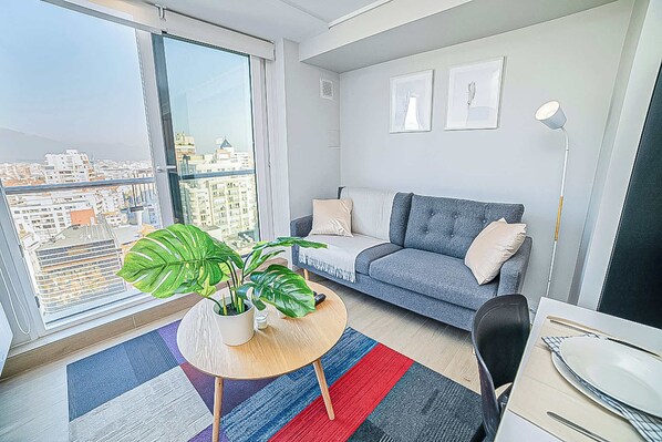 A modern living room with a grey sofa, a small round table with a plant, and a dining area with a white table and chairs. - Functional and Bright Apartment in Providencia (Santiago)