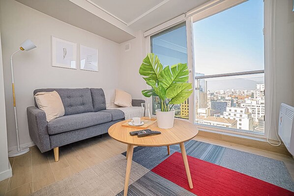 A modern living room with a grey sofa, a round wooden coffee table, a potted plant, and a view of a cityscape through a large window.