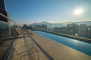 A rooftop pool with a glass railing, sun, and a cityscape in the background.
