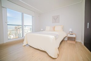 A bedroom with a large window, a bed, a nightstand, and a wall-mounted artwork.