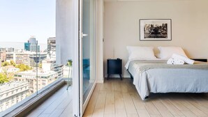 Een slaapkamer met een bed, nachtkastje, een ingelijste foto aan de muur en stadszicht door een groot raam.