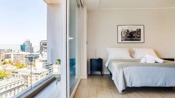 A bedroom with a bed, a nightstand, a framed picture on the wall, and a city view through a large window.