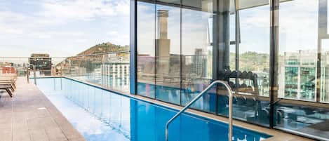 A rooftop pool with clear glass walls, a wooden deck, and a city view.