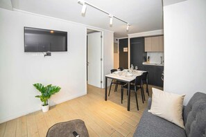 Una sala de estar moderna con un televisor de pantalla plana, una zona de comedor con una mesa preparada para cuatro personas, un sofá gris y una planta.