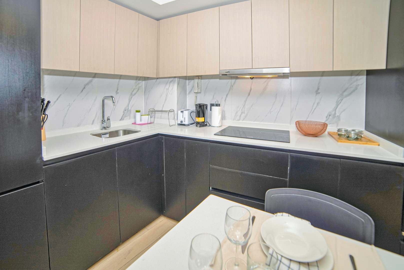 Een kleine keuken met donkere kasten, een wit aanrechtblad, een spoelbak en een eethoek met een tafel gedekt voor twee personen.
