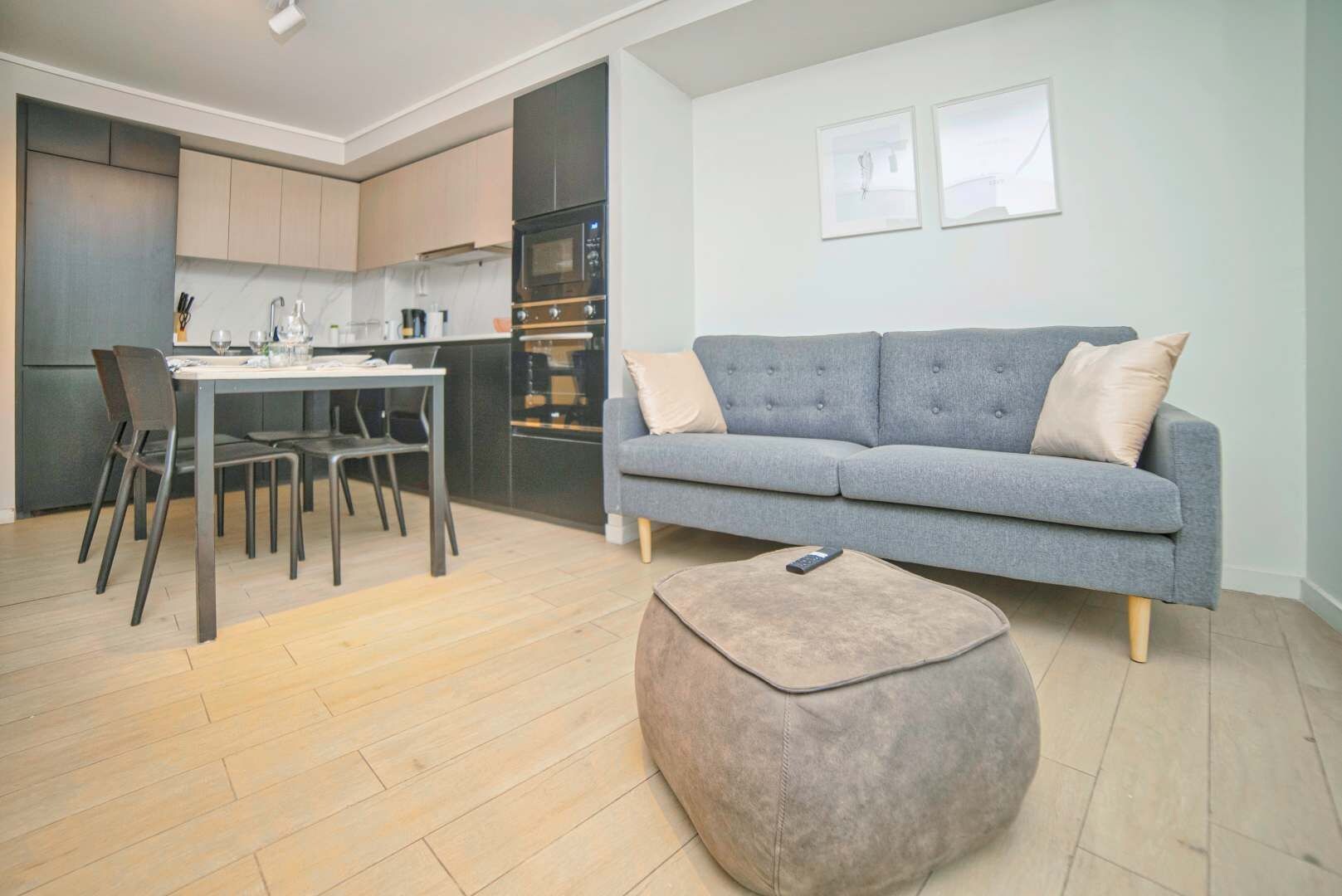Een moderne woonkamer met een grijze bank, een eettafel met stoelen en een keuken op de achtergrond.