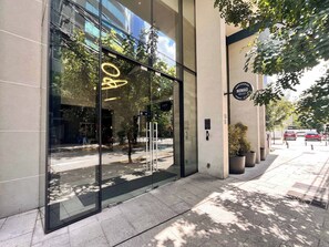 A modern building with large glass windows reflecting trees and a clear sky. There is a sign for 'Arrendadores Nomad' and a street sign indicating a one-way street. - Close to everything in Providencia 2BR2BA Apt (Santiago)