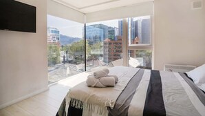 A modern bedroom with a large bed, a flat-screen TV, and a city view through large windows.