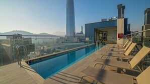 Una piscina en la azotea con sillones de descanso, barandilla de cristal y vista a un rascacielos al fondo.