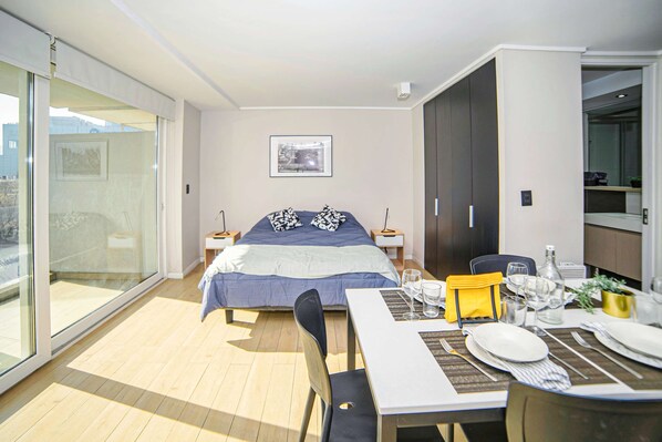Habitación de hotel moderna con una cama grande, una mesa de comedor para dos personas, una puerta corredera que da a un balcón y un cuadro en la pared.