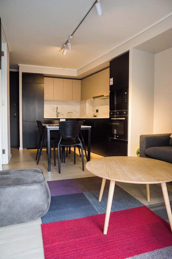 A modern kitchen with black appliances, a dining area with a table and chairs, and a gray sofa. - Modern 1BR Flat Prime Location Luxury Living (Santiago)