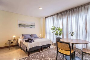 A hotel room with a bed, bedside tables, a desk, a chair, a table, and a window with curtains.