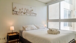 A hotel room with a bed, a nightstand with a lamp, a wall-mounted artwork, and a large window. - Modern Chic Oasis: Sophisticated Las Condes Stay (Las Condes)