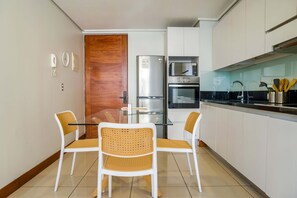 A modern kitchen with white cabinets, a stainless steel refrigerator, an oven, a microwave, and a glass dining table with four chairs.