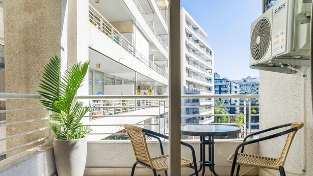 A balcony with a potted plant, two wicker chairs, a table, and a wall-mounted air conditioning unit. - Modern Chic Oasis: Sophisticated Las Condes Stay (Las Condes)