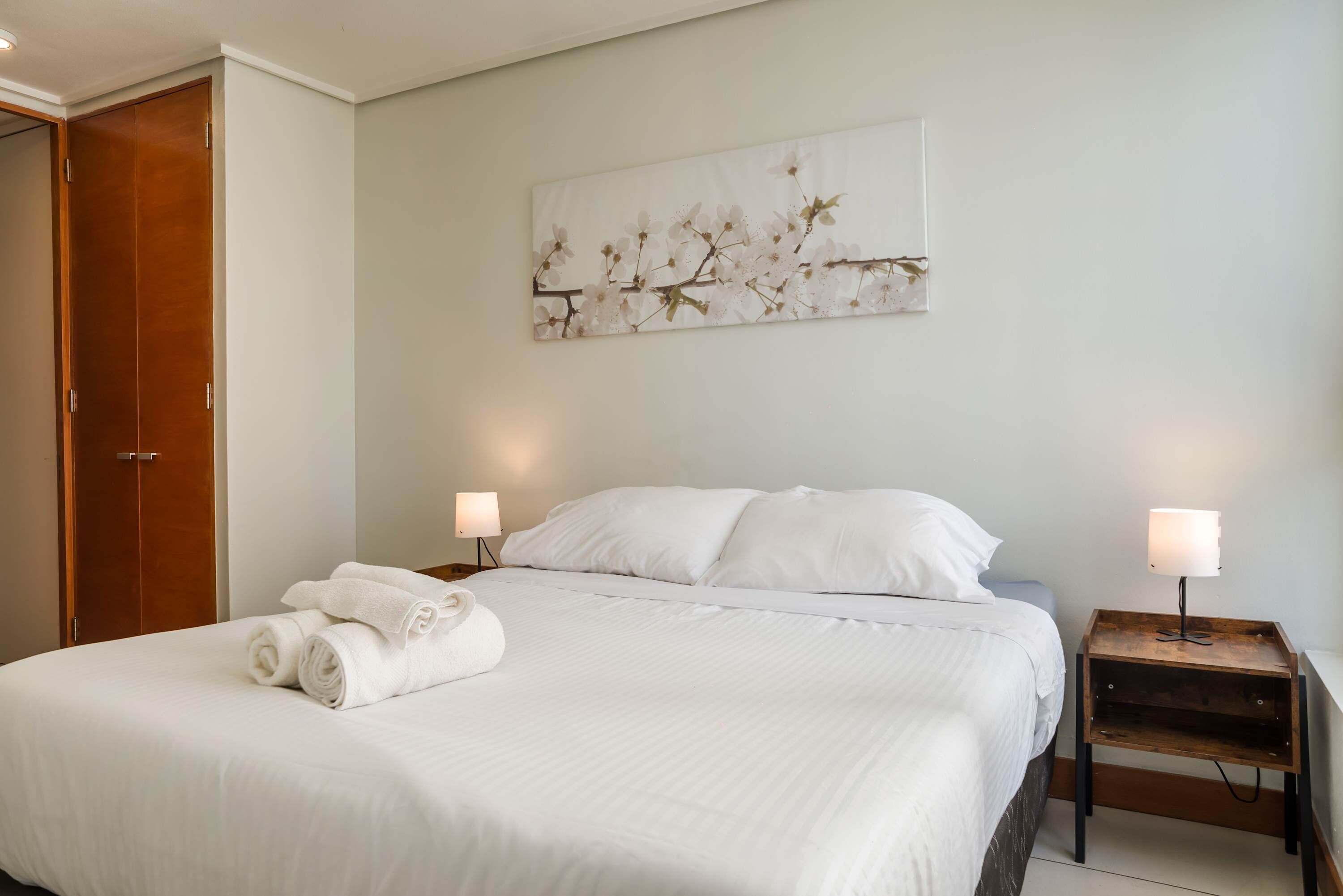 Una cama bien hecha con ropa blanca, una mesita de noche de madera con una lámpara y una obra de arte floral en la pared.
